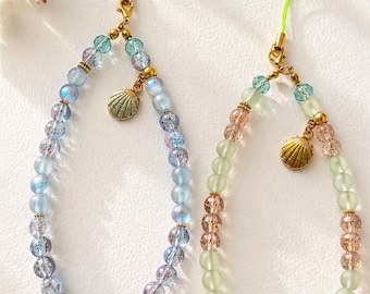 May include: Two beaded phone charms featuring round glass beads in shades of blue, purple, green, and peach. Each charm includes a gold-toned metal seashell pendant, small gold spacer beads, and a thin nylon loop for attachment to a mobile device.