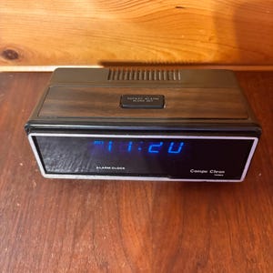 May include: A vintage Compu Chron alarm clock with a black and wood-grain finish. The digital display shows the time in blue. The clock has a rectangular shape with a silver frame and the words "Alarm Clock" and "Compu Chron" printed on the front.