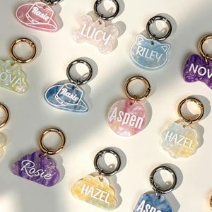 Pode incluir: Uma coleção de chaveiros de acrílico personalizados em várias formas, incluindo nuvens, ossos, círculos e chapéus de cowboy. Cada peça apresenta um acabamento marmorizado e brilhante em cores como rosa, azul, roxo e amarelo.
