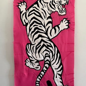 May include: A rectangular fabric wall hanging featuring a pieced quilt design of a white tiger with black stripes. The tiger is depicted in a dynamic, pouncing pose against a vibrant, solid hot pink background made of multiple fabric segments.