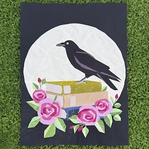 Op de afbeelding: Illustratie van een zwarte kraai die op een stapel boeken zit, tegen een witte maan op een zwarte achtergrond. Roze rozen en groene bladeren sieren de bodem. Het kunstwerk is een quilt- of stoffen ontwerp.