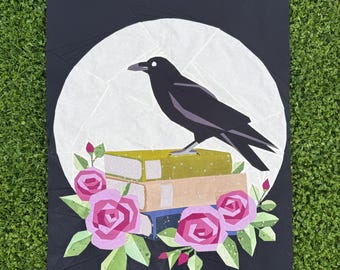 May include: A black crow illustration perched atop a stack of books, set against a white moon on a black background. Pink roses and green leaves adorn the bottom. The artwork is a quilt or fabric design.