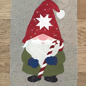 Pode incluir: Um gnomo de tecido com um chapéu vermelho com uma estrela branca e flocos de neve, uma barba branca e um casaco verde. O gnomo segura uma bengala de doces listrada em vermelho e branco. O fundo é cinza com bolinhas brancas e vermelhas.