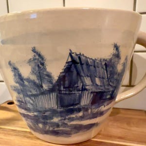 May include: Cream-colored ceramic mixing bowl with a blue landscape design. The bowl features a spout and a large handle. The blue design depicts a barn and trees. The bowl sits on a wooden cutting board.