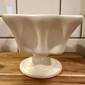 May include: A solid off-white ceramic pedestal bowl featuring a wide, flared rim with a scalloped, undulating edge and a smooth, rounded base. The bowl has a deep, curved interior and a sturdy, central stem connecting the basin to the circular foot.