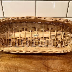 May include: An oval-shaped, light brown woven wicker basket with a sturdy rim and a cross-weave base pattern. A small rectangular white label with the text Crate & Barrel is attached to the center of the interior base of the basket.