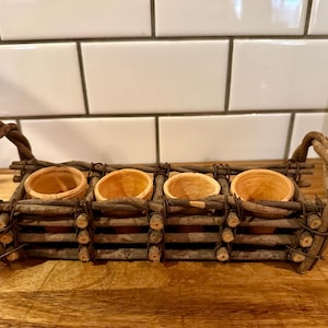 May include: A rectangular holder crafted from dark brown twigs and branches, featuring two arched handles on the ends. The structure contains four small, round, light orange terracotta pots arranged in a single row within the wooden frame.