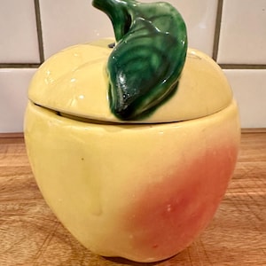 May include: A ceramic lidded jar shaped like an apple. The body is primarily pale yellow with a soft pink blush on one side. A glossy, dark green sculpted leaf and stem form the handle on the lid, which sits atop the rounded apple-shaped base.