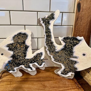 May include: Two fish-shaped decorative objects made of off-white material with a textured surface. Each fish features a central geode-like inlay filled with black, gold, and white granular details, and a single dark dot representing an eye.