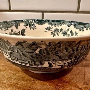 May include: A cream-colored ceramic bowl with two handles, featuring a dark green transferware pattern of a rural landscape with thatched cottages and figures. The interior rim is decorated with a detailed floral and scrollwork border design.