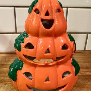 May include: A ceramic jack-o'-lantern stack featuring three orange pumpkins with carved smiling faces and green leaf accents. The hollow structure has cutouts for eyes, noses, and mouths, with a brown stem at the top of the uppermost pumpkin.