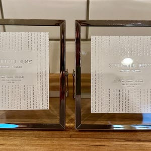 May include: Two square picture frames with polished silver-tone metal borders and clear glass panels. Each frame contains a white insert featuring a vertical dashed pattern and the printed text Sheffield Home Picture Frames Fine Home Decor.