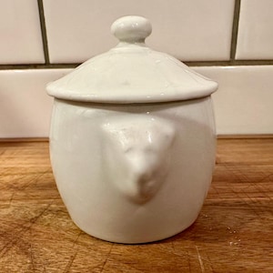 May include: A white ceramic sugar bowl with a matching lid featuring a knob handle. The bowl has a rounded body and a molded relief detail on the side that resembles an animal head. The surface is smooth and glossy with a clean, solid white finish.