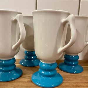May include: A set of four ceramic mugs, each featuring a smooth white cylindrical body with a curved handle and a distinct, tiered pedestal base finished in a glossy, vibrant turquoise blue glaze, arranged in a group on a wooden surface.