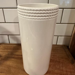 May include: A tall, cylindrical white ceramic vase featuring a smooth, glossy finish. The upper rim of the vase is decorated with a textured, raised rope-like pattern that encircles the entire circumference of the opening, adding detail.