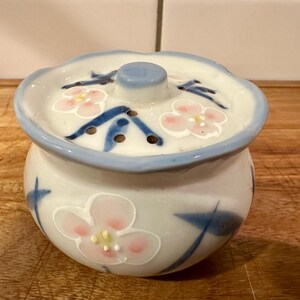 May include: A small, round, white ceramic jar with a matching lid featuring a scalloped edge. The jar and lid are decorated with hand-painted pink flowers with yellow centers and blue leaves. The lid includes a central knob and small holes.