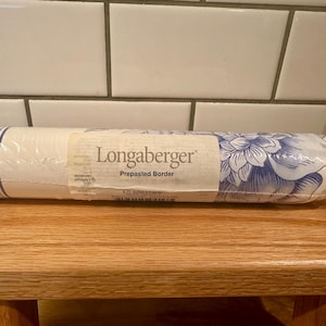 May include: A rolled-up Longaberger prepasted border with a floral design in blue and white. The border is wrapped in clear plastic and rests on a wooden surface. The text "Longaberger" is visible on the label.