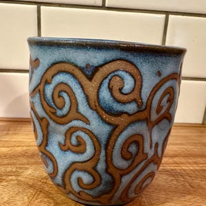 May include: A ceramic mug featuring a light blue glaze with a textured, dark brown swirling vine pattern. The mug has a rounded, tapered shape with a smooth rim and a visible clay texture throughout the surface, showcasing a handcrafted appearance.