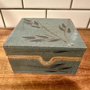 May include: A rectangular ceramic box with a speckled sage green glaze and a dark brown painted leaf motif on the lid and front panel. The box features a notched cutout on the front edge for opening and has a visible raw, unglazed clay rim.