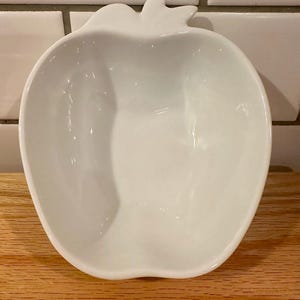 May include: A white ceramic apple-shaped bowl. The bowl has a smooth, glossy finish and a stem detail at the top. It sits on a wooden surface, with white tile in the background.