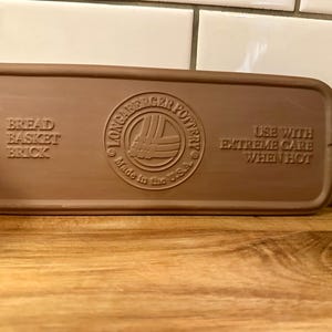 May include: A rectangular brown ceramic bread basket brick. The brick has rounded edges and embossed text that reads "Bread Basket Brick" and "Use with Extreme Care When Hot". The Longacre Pottery logo is also visible.