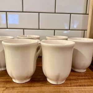 May include: A set of five off-white ceramic mugs with a smooth, glossy finish. Each mug features a rounded body with subtle vertical fluting along the base and a curved handle on the side. The mugs are arranged in a group, showcasing their uniform design.