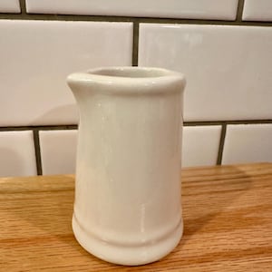 May include: A small, off-white ceramic creamer pitcher with a rounded base and a spout. The creamer is on a wooden surface, with white subway tile in the background.