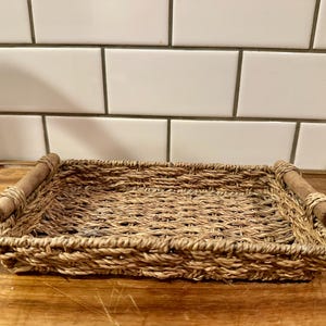 May include: A rectangular woven seagrass tray featuring a natural tan color and a textured, braided pattern. The tray includes two cylindrical wooden handles attached to the shorter sides with woven seagrass accents, providing a sturdy grip.