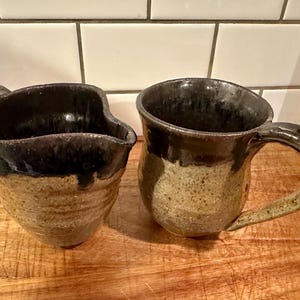 May include: A ceramic creamer pitcher and a matching mug featuring a two-tone design. The lower sections have a speckled tan finish, while the upper rims are coated in a dark, glossy black glaze that drips slightly down the textured surface.