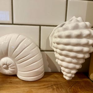 May include: Two white ceramic seashell sculptures. One is a nautilus shape, and the other is a conch shell. Both are glossy and sit on a wooden surface against a white tile background. Decorative coastal home decor.