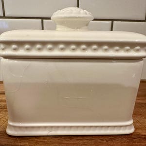 May include: A rectangular white ceramic butter dish with a lid featuring a decorative beaded border along the top edge and a rounded knob handle on top. The base of the container also has a subtle embossed line detail along the bottom edge.