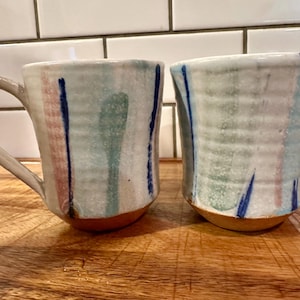 May include: Two ceramic mugs with a textured, ribbed surface and a white base glaze. Each mug features vertical brushstrokes in shades of blue, green, and pink. The mugs have a curved handle and a natural, unglazed terracotta-colored base.