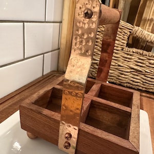 May include: A square wooden caddy with four compartments and a central handle. The handle is made of hammered copper metal, secured with visible screws to the wooden frame. The caddy stands on four small, rounded copper-colored feet.