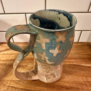 May include: A ceramic mug featuring a two-tone design with a blue-green upper section and a light beige lower section. The surface is decorated with a repeating pattern of tan, cross-shaped marks. It has a large, curved handle that extends downward.
