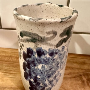 May include: A speckled ceramic tumbler with a flared rim. The off-white surface is decorated with blue and green abstract floral designs. The tumbler is approximately 6 inches tall.