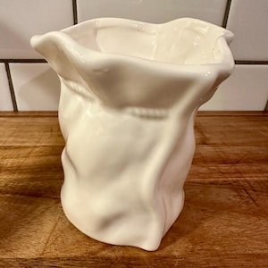 May include: A white ceramic vase shaped like a crumpled paper bag. The vessel features a glossy finish with irregular, folded edges at the top and a textured, creased surface that mimics the appearance of a soft, flexible material gathered at the neck.