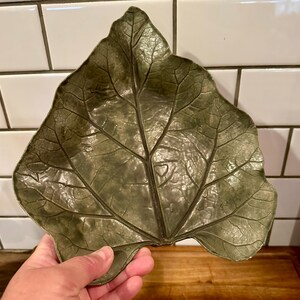 May include: A leaf-shaped decorative dish in a muted olive green color with a glossy finish. The surface features a detailed, raised vein pattern that mimics the natural structure of a broad leaf, with a pointed tip and a wide, lobed base.