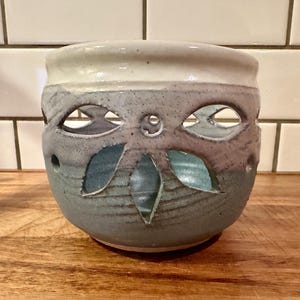 May include: A ceramic candle holder with a rounded shape, featuring a cut-out floral pattern around the center. The glaze transitions from a creamy white at the top rim to a speckled light blue and grey finish on the lower body of the vessel.