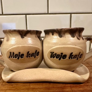 May include: Two beige ceramic mugs with dark brown drip glaze at the rims, each featuring an oval plaque with embossed text. The left mug reads Moje kafe and the right mug reads Tvoje kafe. The mugs sit on a matching beige ceramic tray.