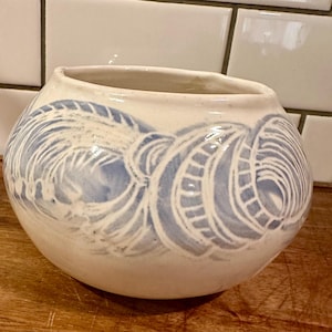 May include: A round, off-white ceramic bowl featuring a hand-painted blue pattern of swirling, circular, and linear motifs. The vessel has a wide, open mouth and a smooth, glossy finish, showcasing a distinct contrast between the blue and white.