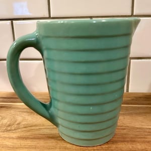May include: A light teal ceramic pitcher featuring a series of horizontal ridges along the exterior body. The pitcher has a curved handle on the left side and a small pouring spout at the top rim, with a smooth, glossy finish throughout the piece.