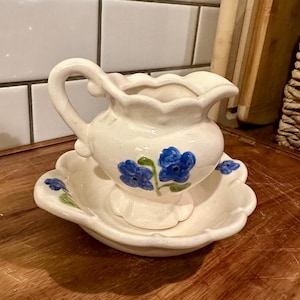 May include: A cream-colored ceramic creamer pitcher and matching saucer set featuring a scalloped edge design. The pitcher has a curved handle and a pouring spout, decorated with hand-painted blue flowers and green stems on the exterior surface.