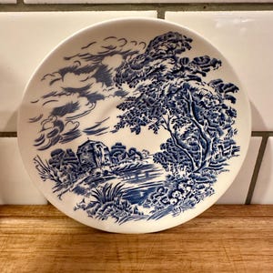May include: A round white ceramic saucer featuring a blue transferware pattern of a landscape. The design depicts a large tree on the right, a small building in the distance, and swirling clouds in the sky, all rendered in detailed blue ink.