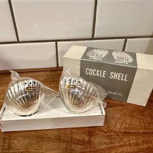 May include: Two silver-toned metal salt and pepper shakers shaped like cockle shells, partially wrapped in clear plastic. They are presented in a white cardboard box with a gray label reading COCKLE SHELL SHAKERS and illustrations of shells.
