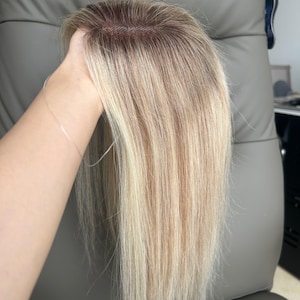 Puede incluir: Un postizo de cabello liso en tonos rubios variados con una raíz castaña más oscura. La pieza incluye una base de encaje visible en la coronilla, diseñada para integrarse con el cabello natural, mostrando una textura suave y sedosa.