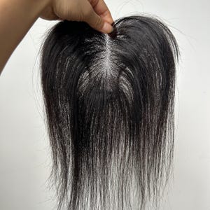 May include: A dark brown hair topper with a natural-looking part. The hairpiece appears to be made of real hair, with strands of varying lengths. The hair is held up by a hand against a white background.