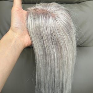 Pode incluir: Um aplique de cabelo com fios lisos em tom cinza-prateado claro e uma base de monofilamento visível no topo. O cabelo é liso e sedoso, apresentando uma risca de aspeto natural onde as madeixas se unem ao material da base cor de pele.