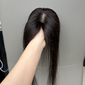 May include: A dark brown hair topper with long, straight strands. The hairpiece is held up, showcasing the natural-looking part and the texture of the hair. The hairpiece appears to be made of human hair.
