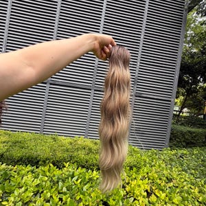 以下が含まれることがあります： ウェーブのかかった質感とオンブレカラーのグラデーションが特徴の長いヘアエクステンション。上部は暗いブラウンの色合いで、毛先に向かって徐々に明るいアッシュブロンドのトーンへと変化しています。