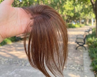 Puede incluir: Un postizo de cabello con mechones lisos de color marrón medio y sutiles tonos rojizos. La base está construida con un material de malla fina similar a la piel para su fijación, mostrando una raya de aspecto natural y textura suave.